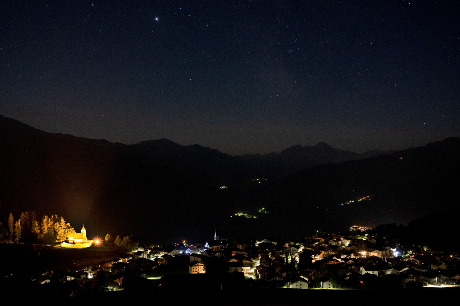 Nachtaufnahme des Südhimmels und des Dorfs Falera in den Schweizer Alpen. Gut zu sehen ist die Lichtverschmutzung durch die Beleuchtung der Kirche.